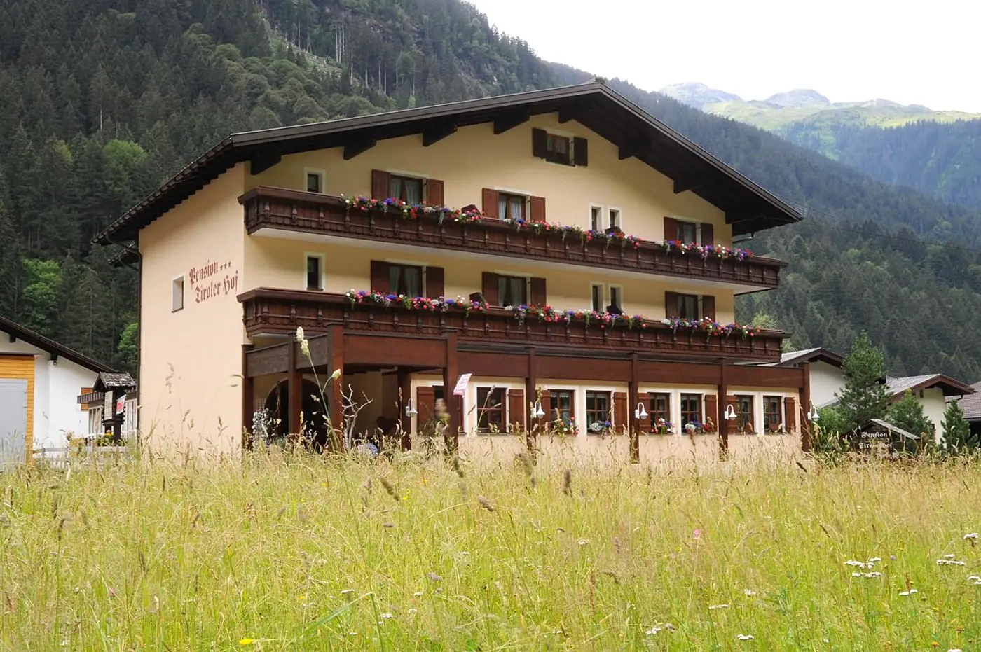 Der Tirolerhof im Sommer, Balkone mit Blumen, davor eine Wiese, dahinter Wald